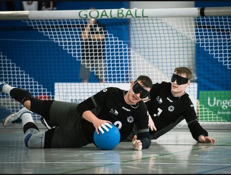 Zwei Spieler liegen in schwarzer Spielkleidung auf dem Boden, beide tragen Dunkelbrillen. Der vordere Spieler hält einen blauen Ball in der Hand. Im Hintergrund ist ein Tor mit der Aufschrift "Goalball"zu sehen.