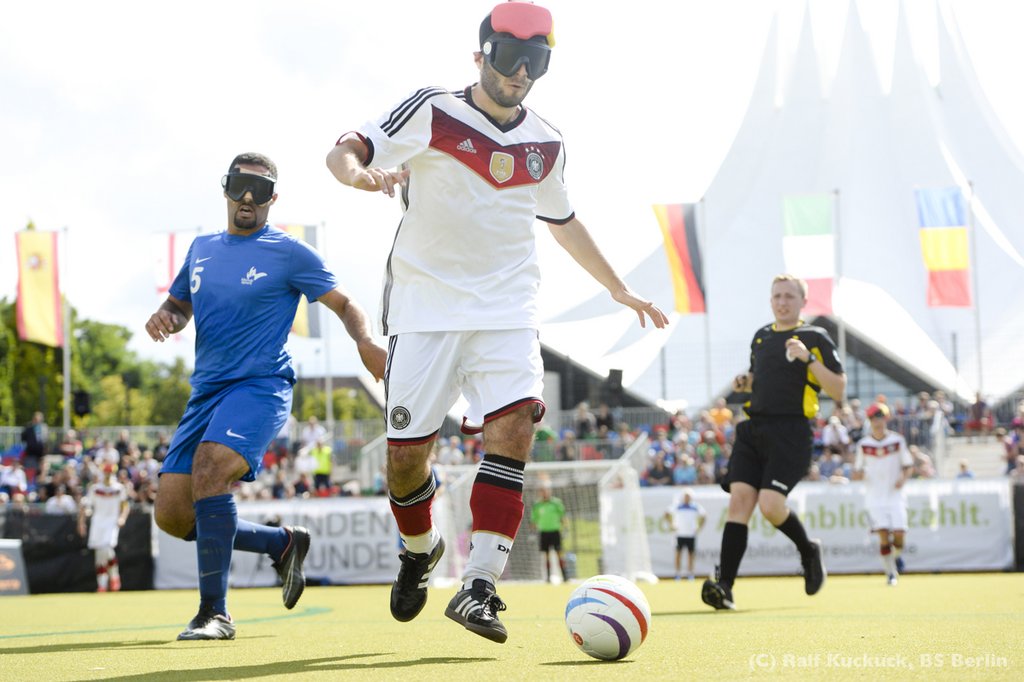 Ein Fußballer im Deutschland-Trikot mit blickdichter Brille führt den Ball mit dem Fuß. Im Hintergrund ein kleines Stadion mit Zuschauern.