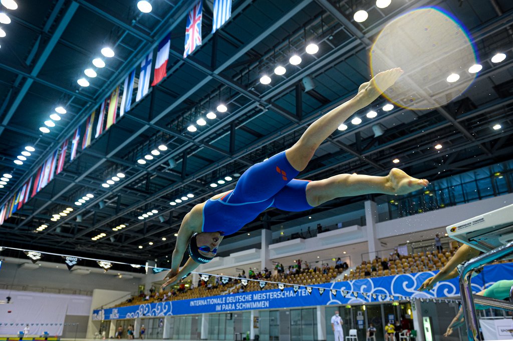 Eine Schwimmerin springt vom Startblock ins Wasser. Parspektive von unten mit Blick unter das Dach der Schwimmhalle.