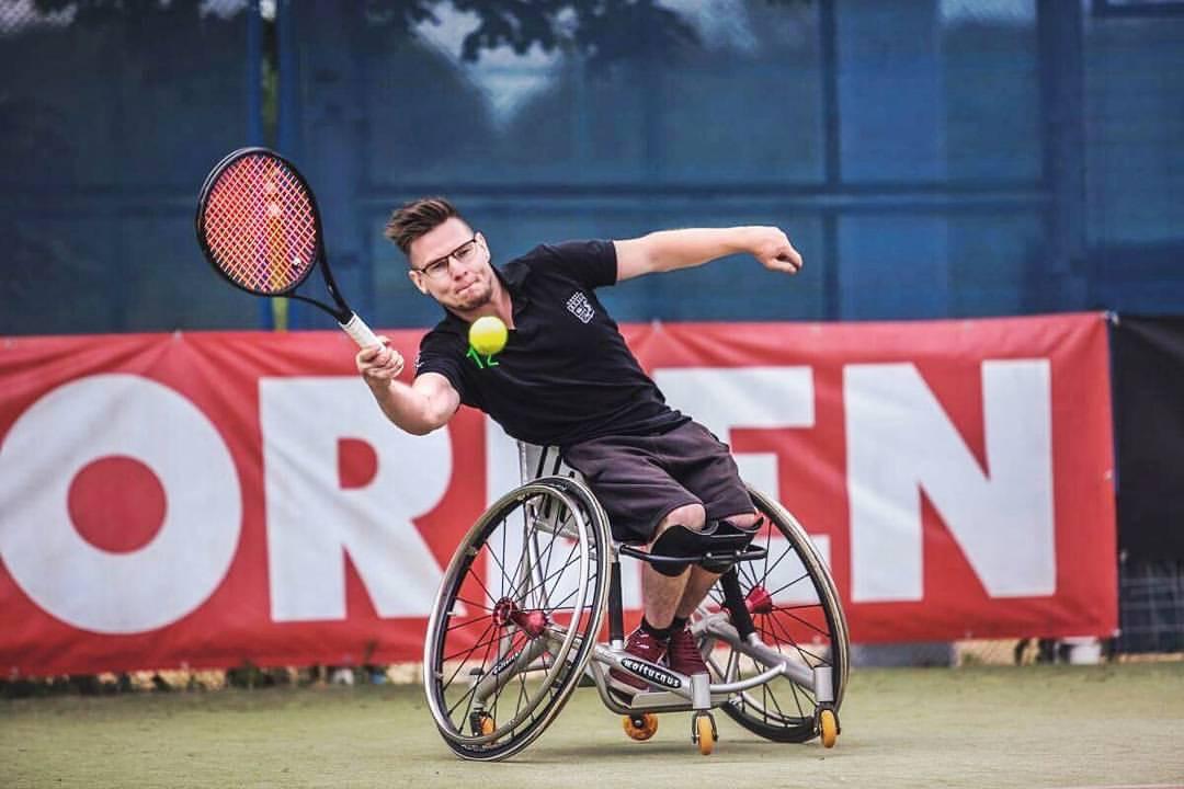 Ein junger Mann spielt im Rollstuhl sitzend Tennis. Mit dem rechten Arm führt er eine Schlagbewegung Richtung Ball aus. Sein Körper ist dabei dynamisch zur rechten Seite geneigt.